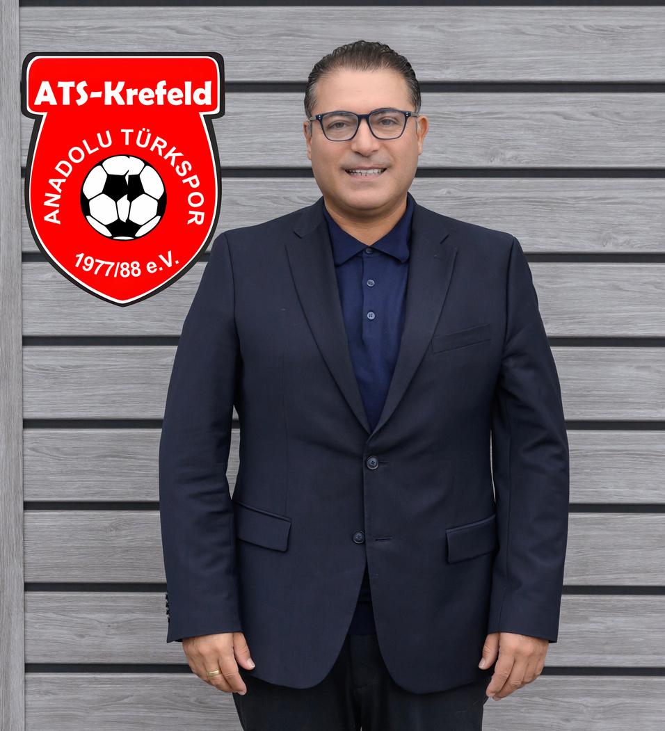 Mann in Anzug mit Brille steht vor grauer Wand, links daneben Vereinslogo von Anadolu Türkspor.